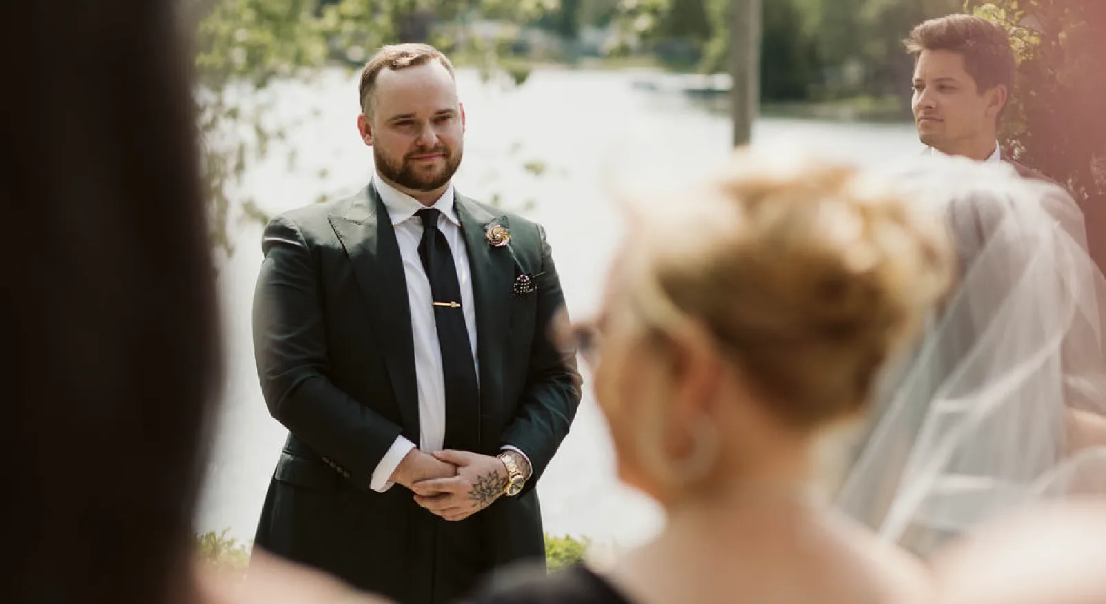 Groom in a dark suit and tie standing outdoors looking at a bride in a veil during a wedding ceremony.