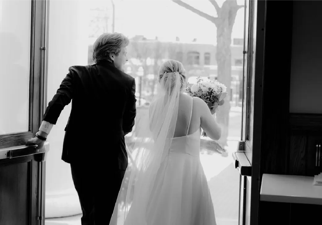 Bride holding a bouquet and a man in a suit opening a door as they exit a building.