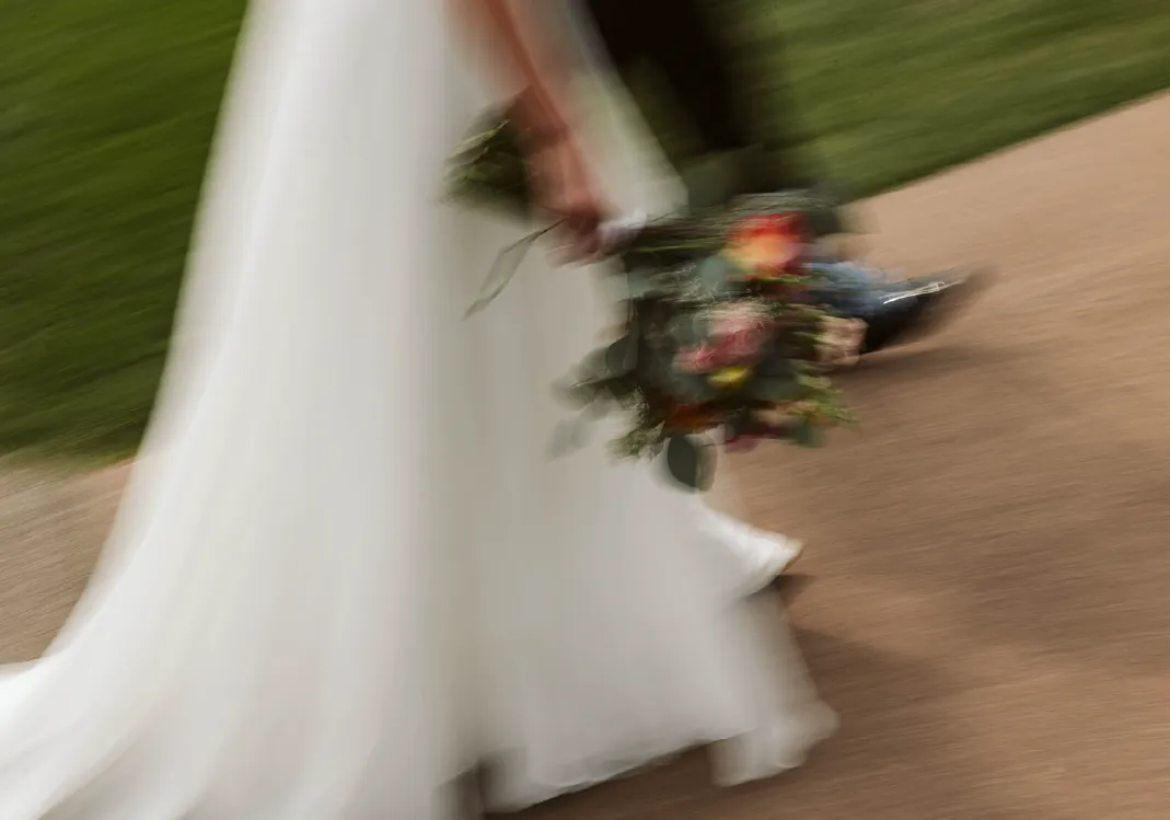Blurred motion image of a person in a flowing white dress holding a bouquet of flowers walking on a pathway next to grass.