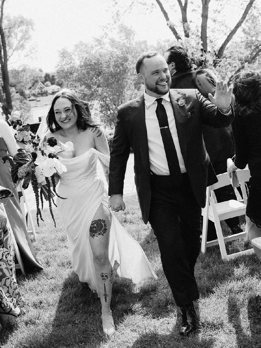 Bride and groom happily walking hand in hand outdoors, bride holding bouquet and showing tattoos on her leg, groom waving to guests.