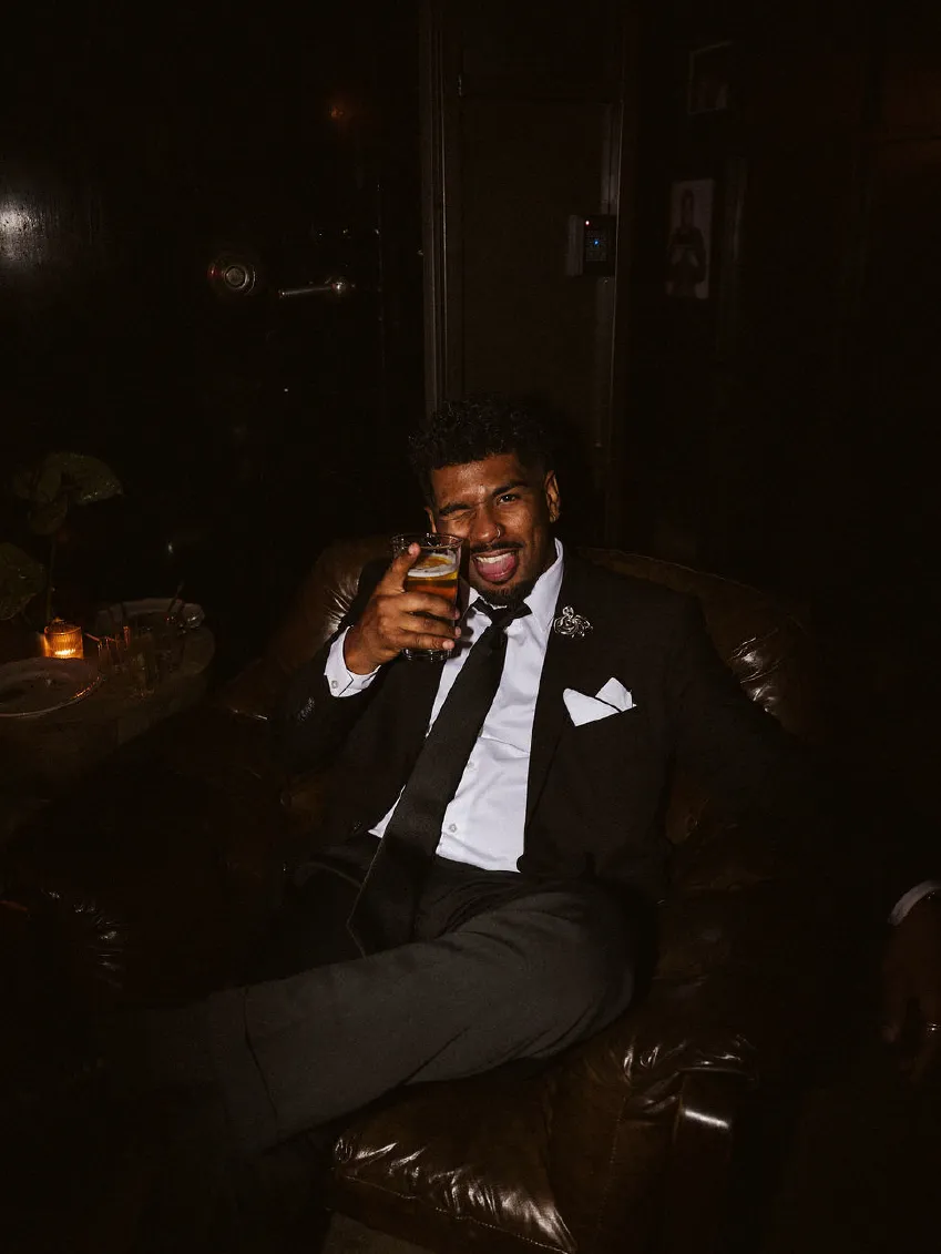Man in black suit sitting in a leather armchair, holding a drink and smiling with one eye closed.