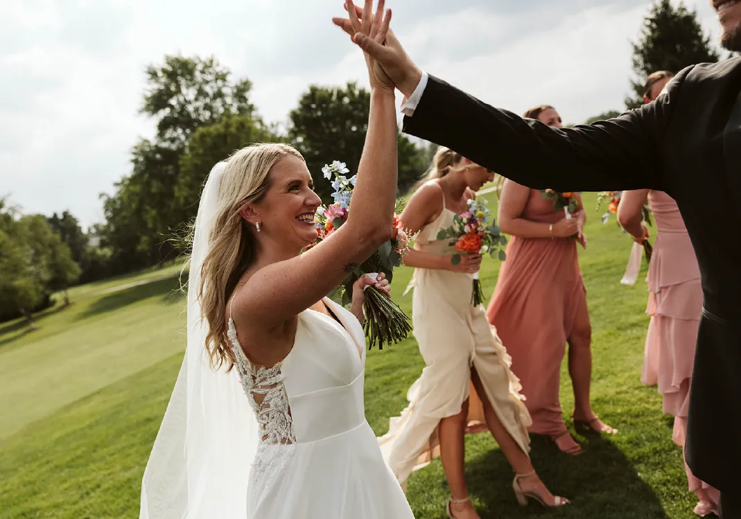 Bride in white dress holding a bouquet and giving a high-five outdoors with bridesmaids in pastel dresses in the background.