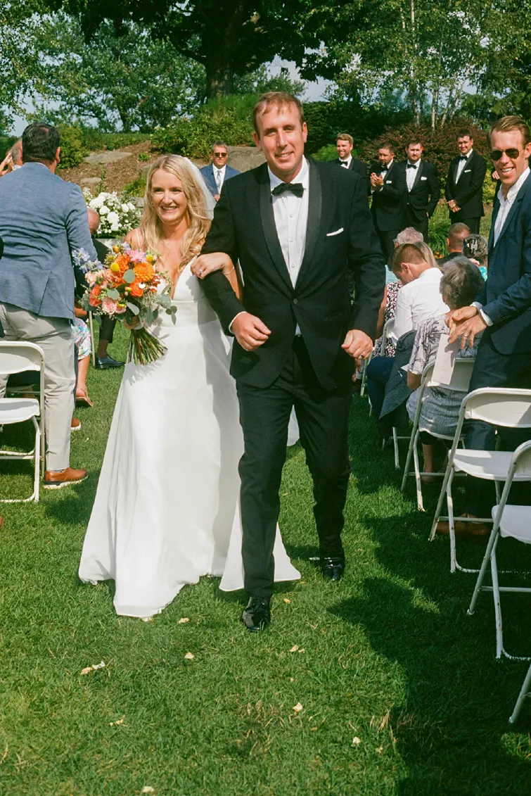 Bride in a white dress holding a colorful bouquet and groom in a black tuxedo walking down a grassy aisle outdoors, surrounded by guests and groomsmen.