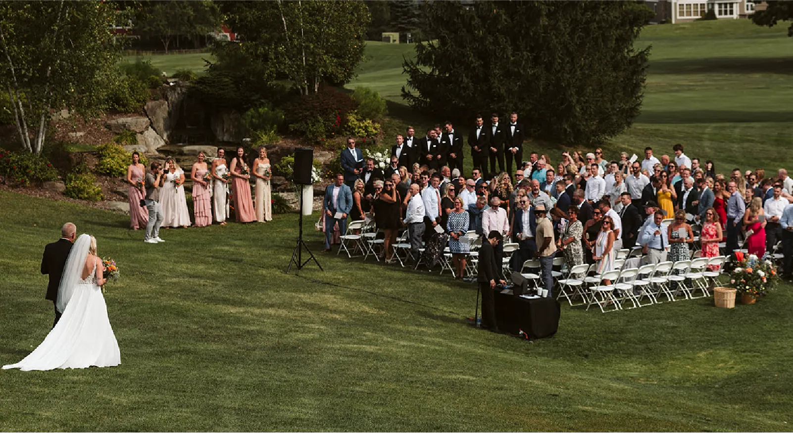 Bride in white gown walking down the grassy aisle with a man towards seated wedding guests and bridesmaids standing nearby.