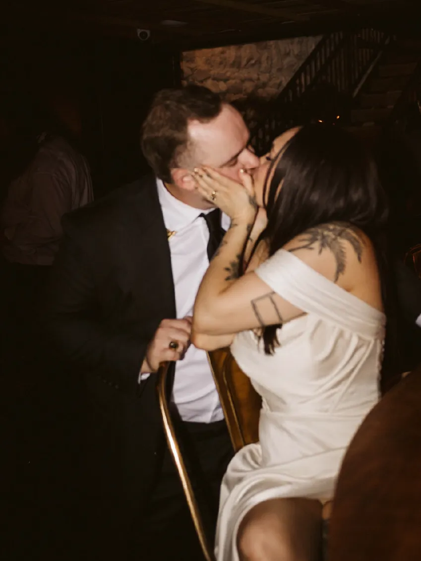 A couple sharing a kiss while seated closely together at an indoor event, the woman wearing a white dress with visible tattoos on her arm and shoulder.