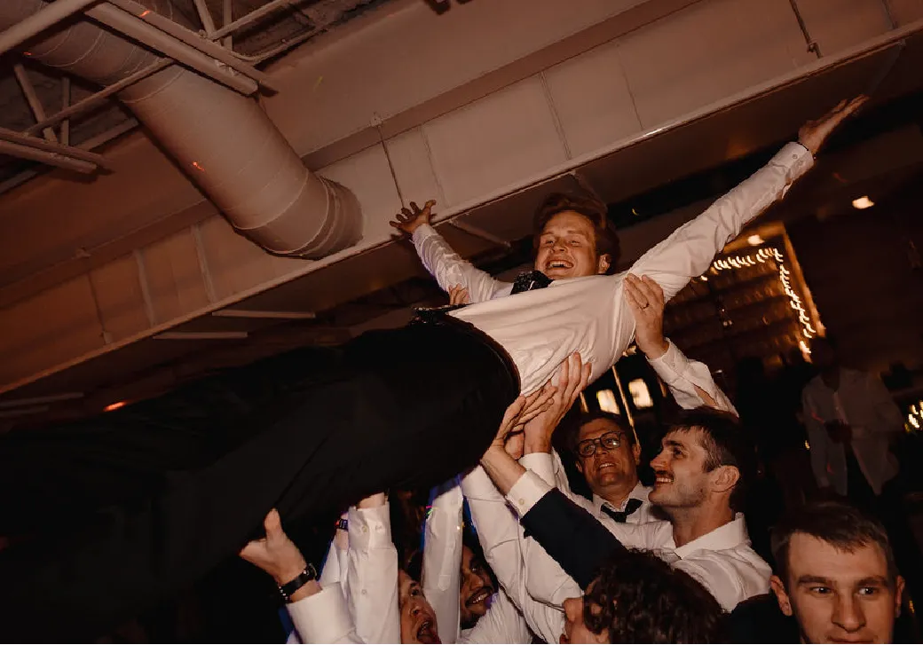 Group of men lifting a smiling man in a white shirt and black pants above their heads indoors at a celebration.