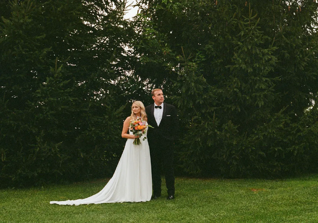 Bride in white gown holding a bouquet standing beside groom in black tuxedo in front of large evergreen trees on green grass.