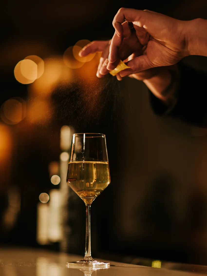 Hand zesting a lemon peel over a glass of white wine with warm bokeh lights in the background.