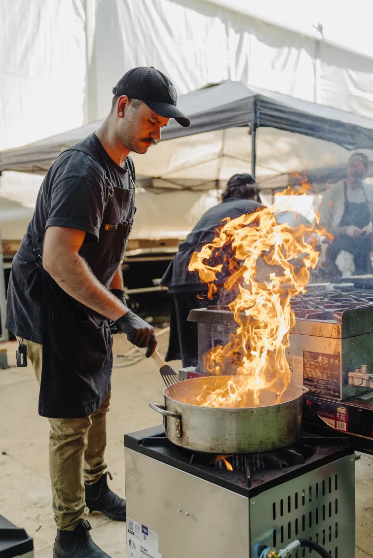 Chef in black apron and cap cooking with a large flame rising from a pan on a gas stove.