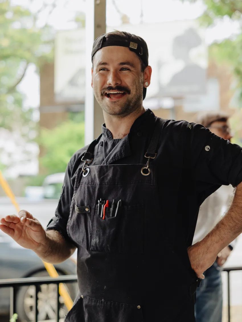 Smiling man in black chef uniform and cap talking with hand raised, standing outdoors.