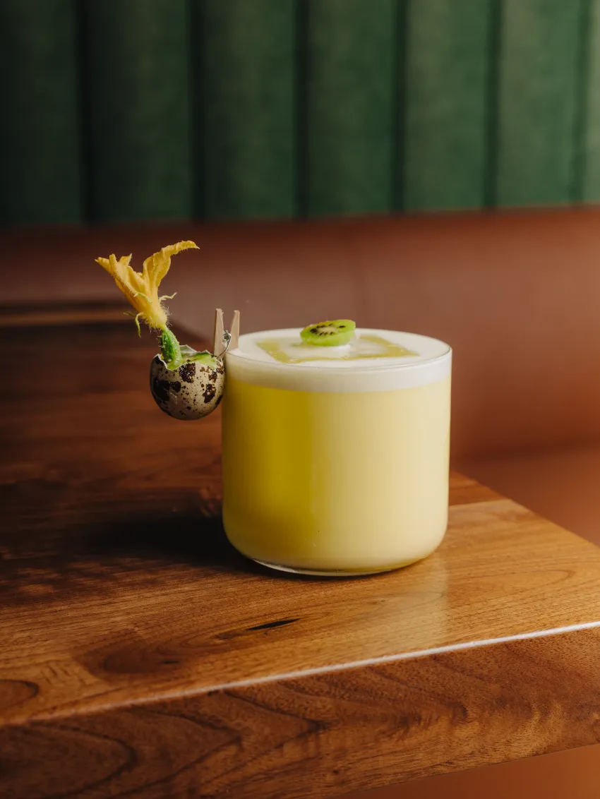 Yellow creamy cocktail in a glass with foam on top, garnished with a small kiwi slice and a flower attached to a speckled quail egg shell clipped to the rim.