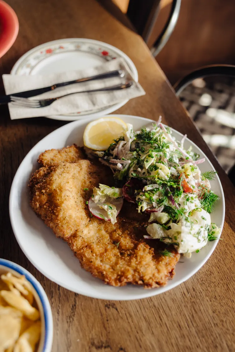 White plate with breaded fried meat cutlet, lemon wedge, and fresh mixed salad with herbs on a wooden table.