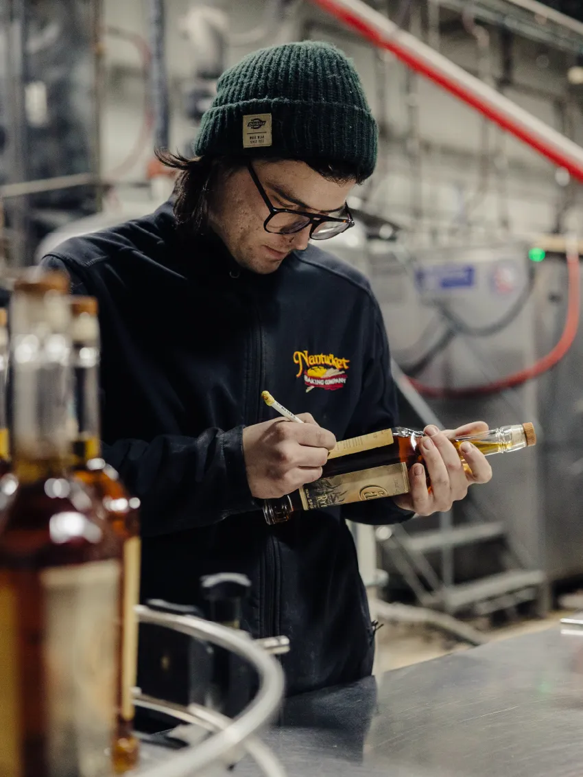 Man wearing a green beanie and glasses labeling a bottle in a distillery or factory setting.