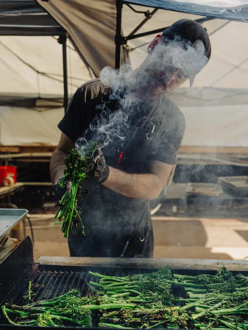 Person wearing black gloves and apron grilling green vegetables on an outdoor grill with smoke rising.
