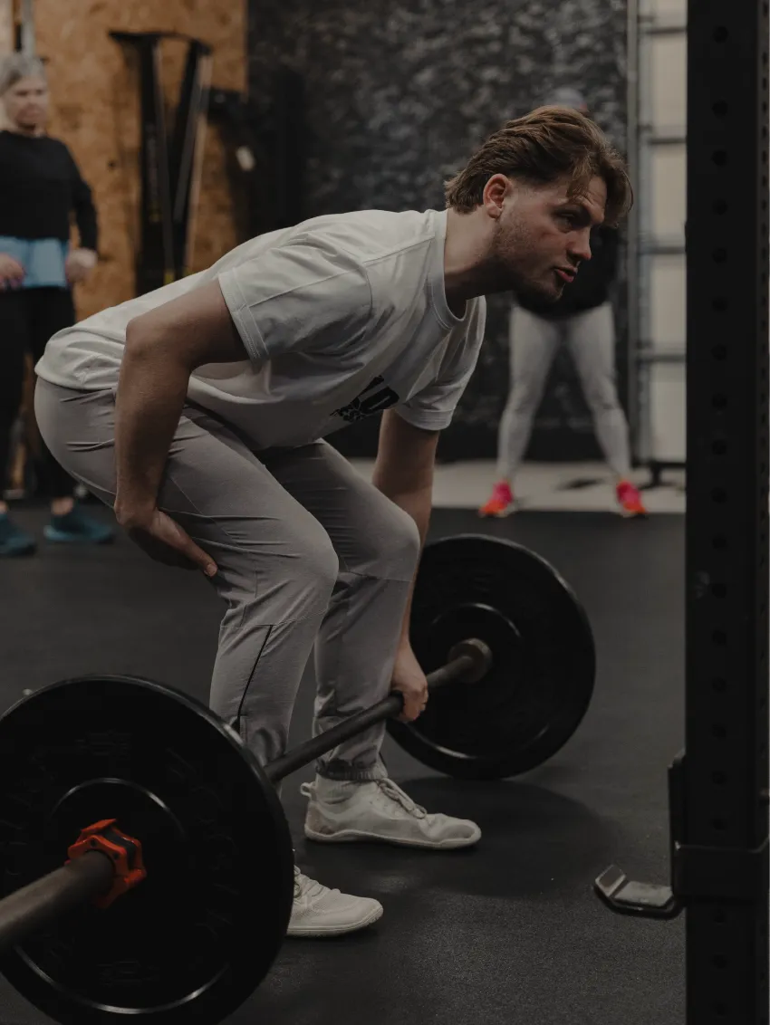 Man in white t-shirt and gray pants lifting a barbell in a gym with two people in background.