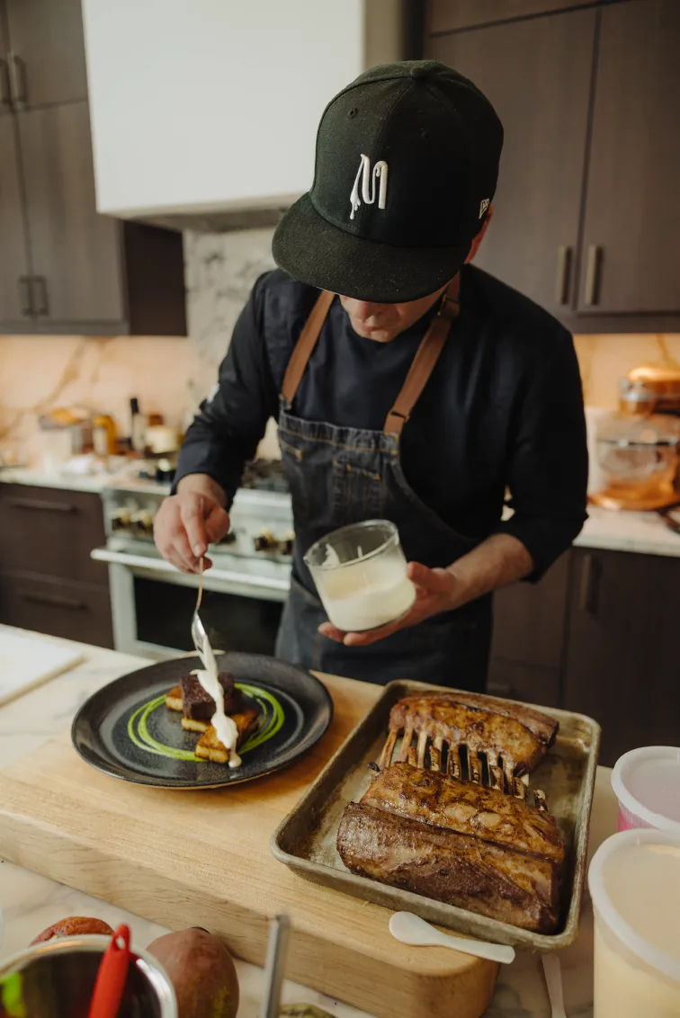 Chef wearing a black cap and apron carefully spooning sauce onto a plated dish in a kitchen with grilled meat on a tray nearby.