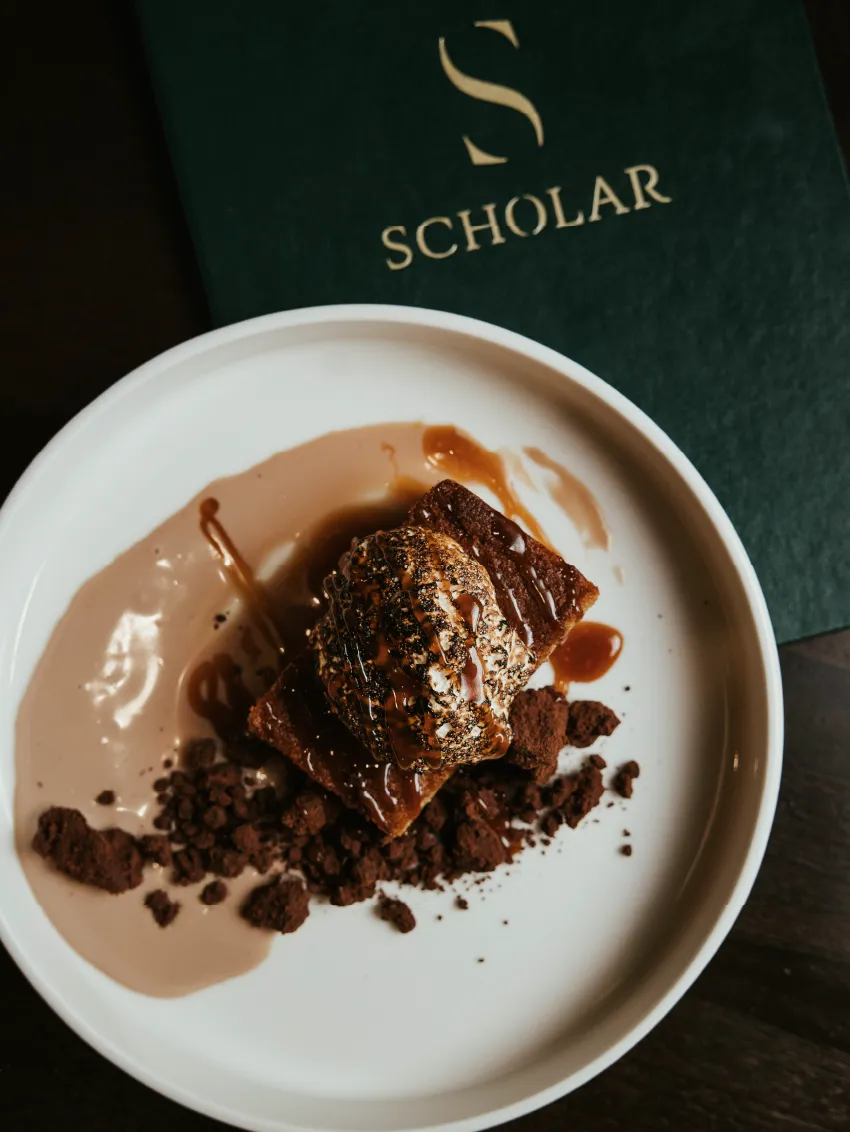 Plate with chocolate dessert topped with cream and chocolate drizzle, served with crumbled chocolate on the side, with a Scholar branded menu in the background.
