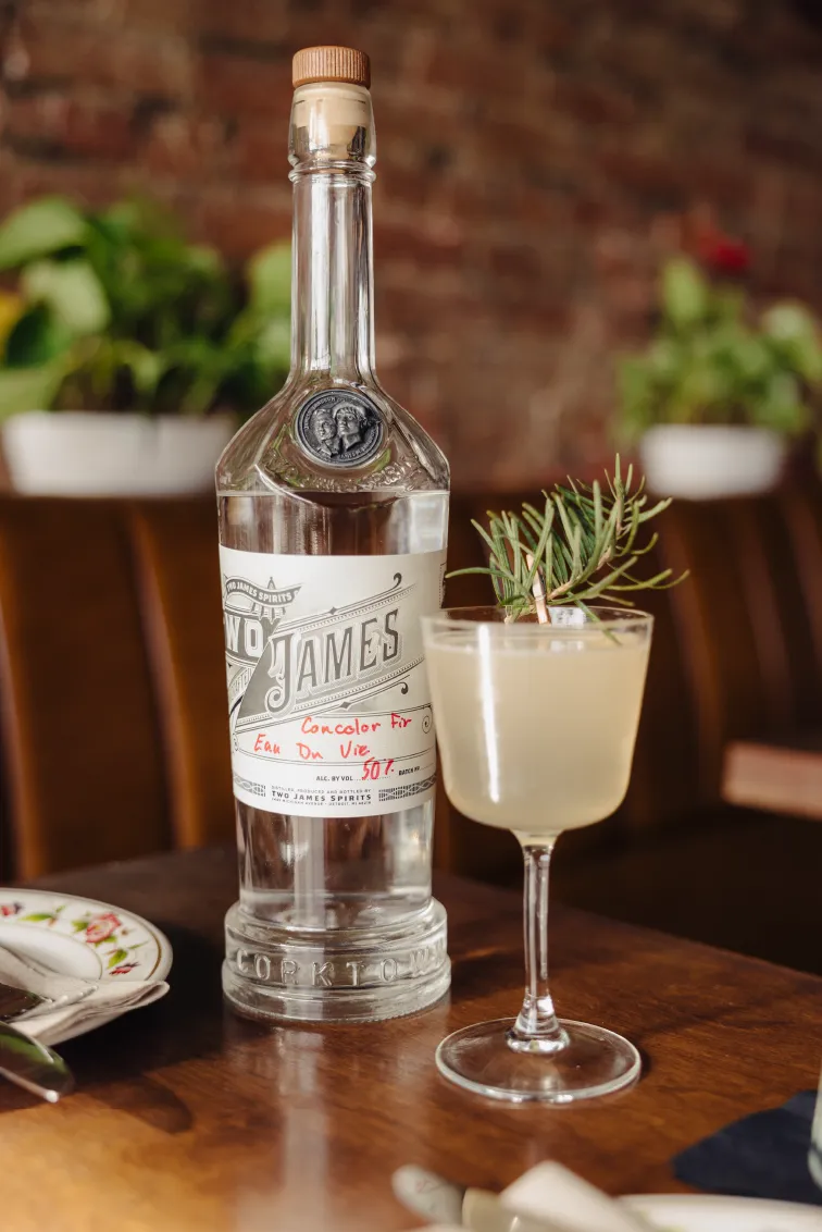 Clear bottle of Two James spirits next to a cocktail glass garnished with a rosemary sprig on a wooden table.