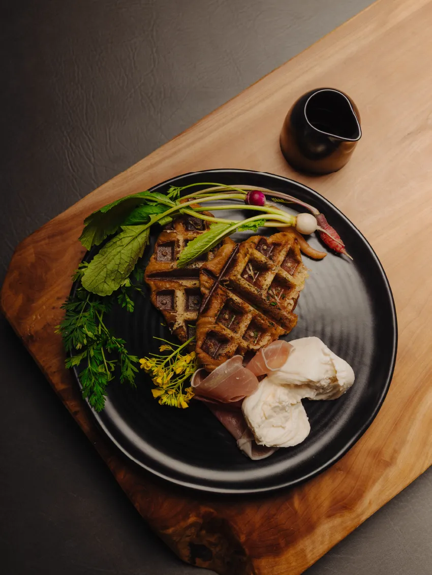 Black plate with two waffles, fresh greens, radishes, slices of prosciutto, and burrata cheese on a wooden board with a small black sauce pitcher.