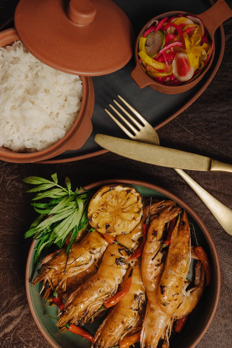 Plate of grilled shrimp with lemon slice and herbs, served with white rice in a clay pot and a side of pickled vegetables, accompanied by gold fork and knife on a dark surface.