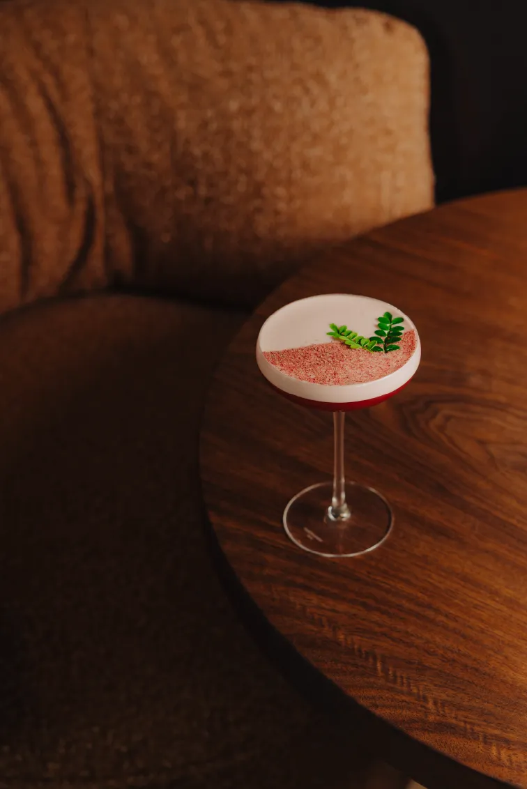 Cocktail with frothy top garnished with green leaves and red powder served in a coupe glass on a wooden table.