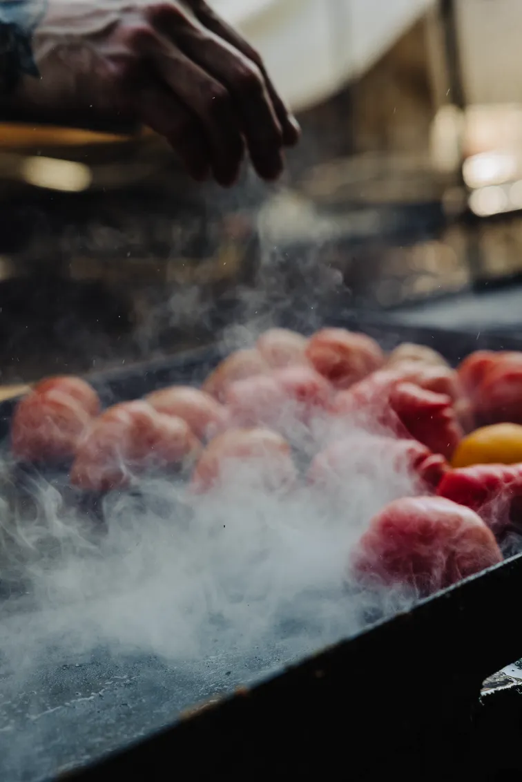 Hand sprinkling seasoning over steaming red tomatoes on a grill.