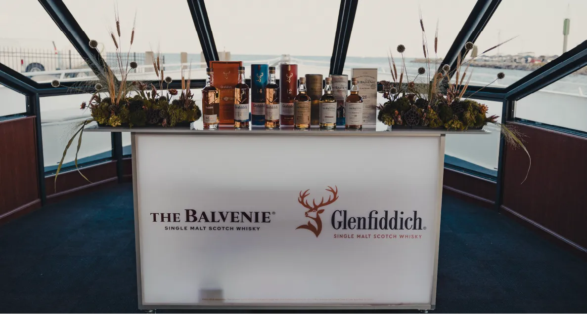Display table with various bottles of The Balvenie and Glenfiddich single malt Scotch whisky inside a glass-walled room overlooking a marina.