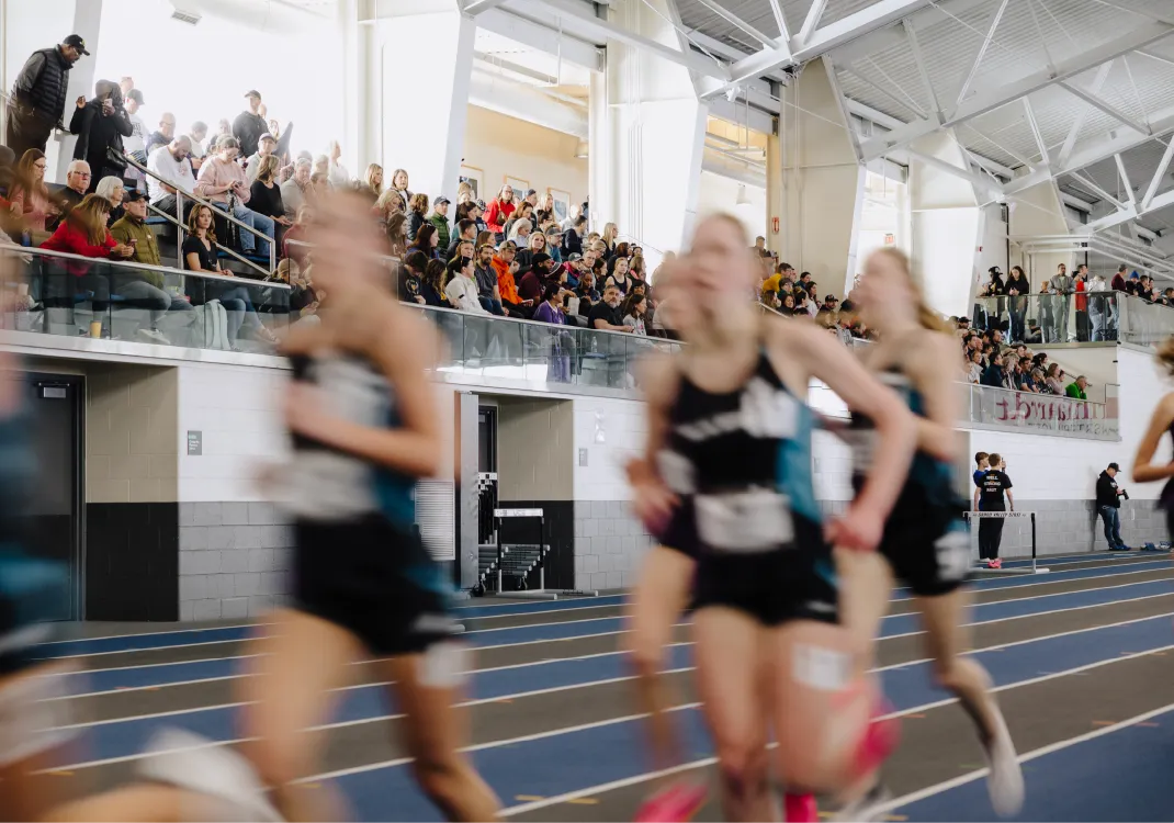 Blurry runners competing on an indoor track with a crowd watching from bleachers.