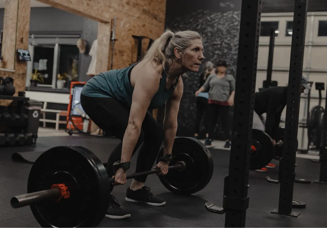 A woman in workout clothes lifting a barbell in a gym with other people exercising in the background.