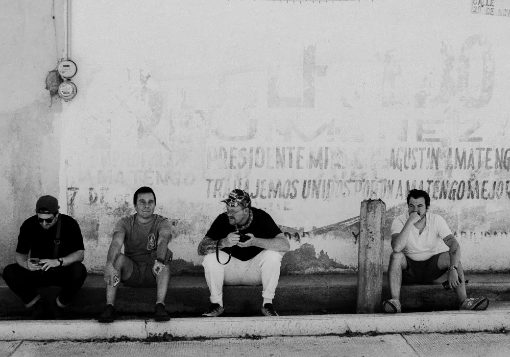 Black and white photo of four men sitting on a curb against a worn wall with faded text.
