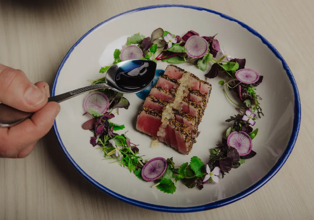Hand holding a spoon with dark sauce above a plate of sliced seared tuna garnished with edible flowers, greens, and purple radish slices arranged in a circular pattern.