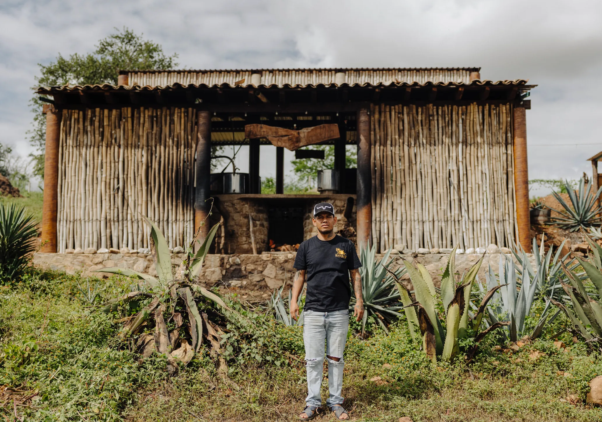 Man wearing a black t-shirt and cap standing in front of a rustic wooden structure surrounded by agave plants.