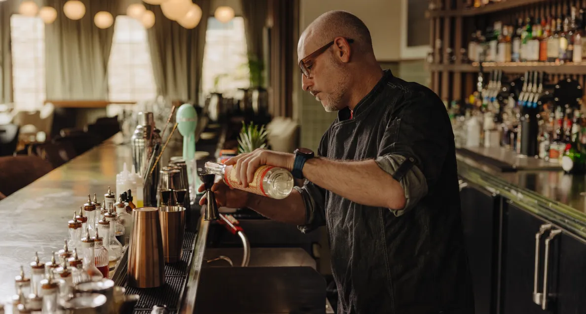 Bartender pouring liquor into a jigger behind a bar with various bottles and tools.