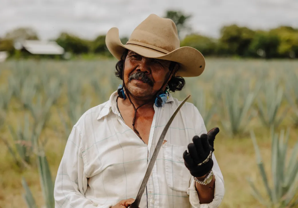 Man wearing a cowboy hat and white shirt holding a machete in an agave field.