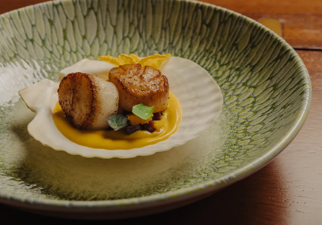 Two seared scallops served on a white shell with yellow sauce and small green leaves in a textured green bowl.