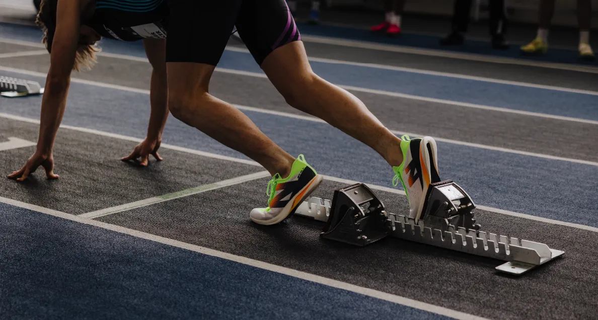 Runner in starting position on indoor track with bright green and white running shoes pressing against starting blocks.