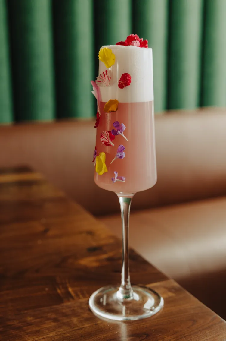 Pink cocktail with white foam in a tall glass, decorated with colorful edible flower petals, on a wooden table.