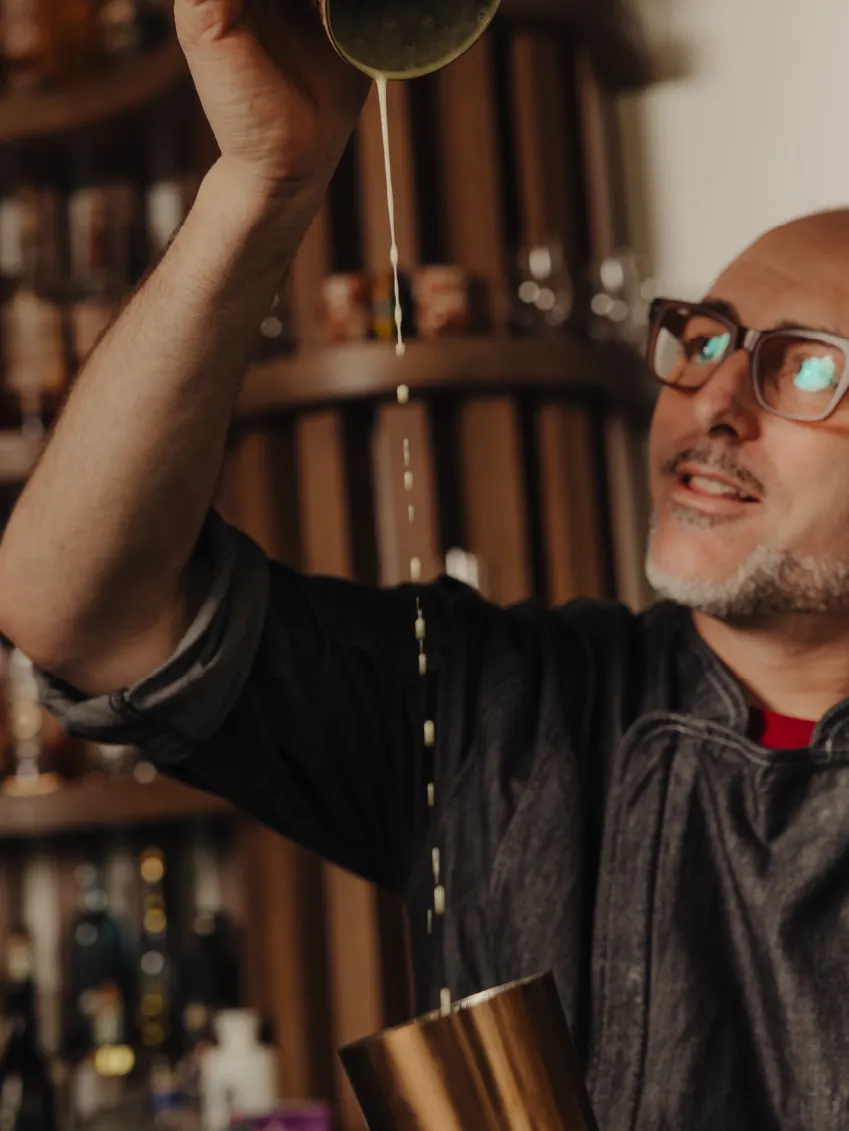 Man with glasses pouring liquid from a shaker into a metal cup in a bar setting.