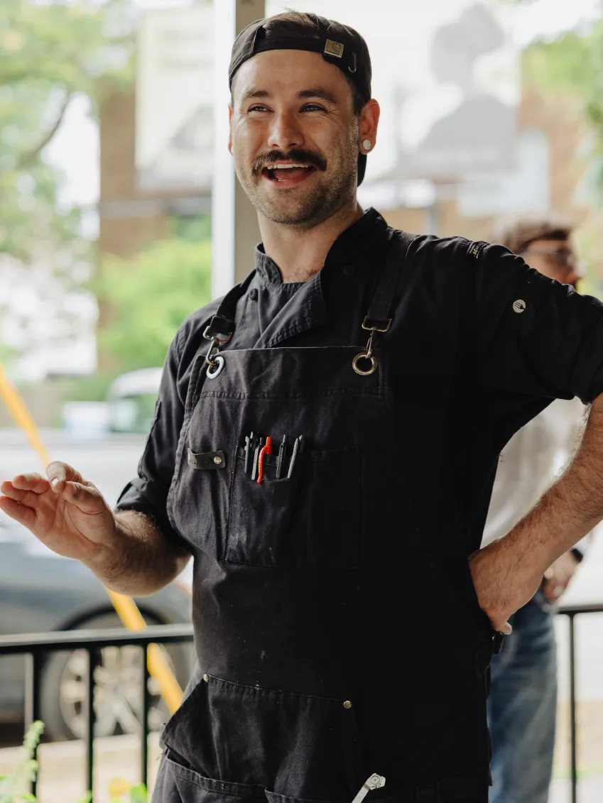 Smiling chef wearing a black cap and apron with utensils in the pocket, gesturing with one hand.