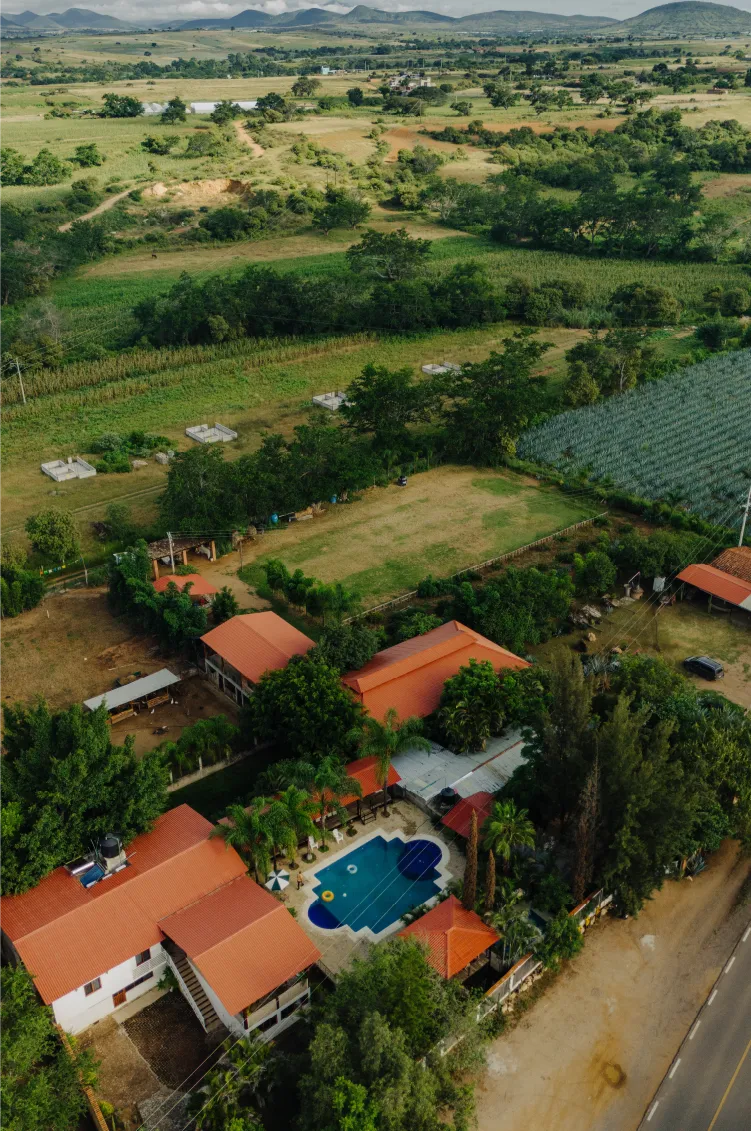 Aerial view of a countryside property with red-roofed buildings, a swimming pool, surrounded by green fields and trees with mountains in the background.