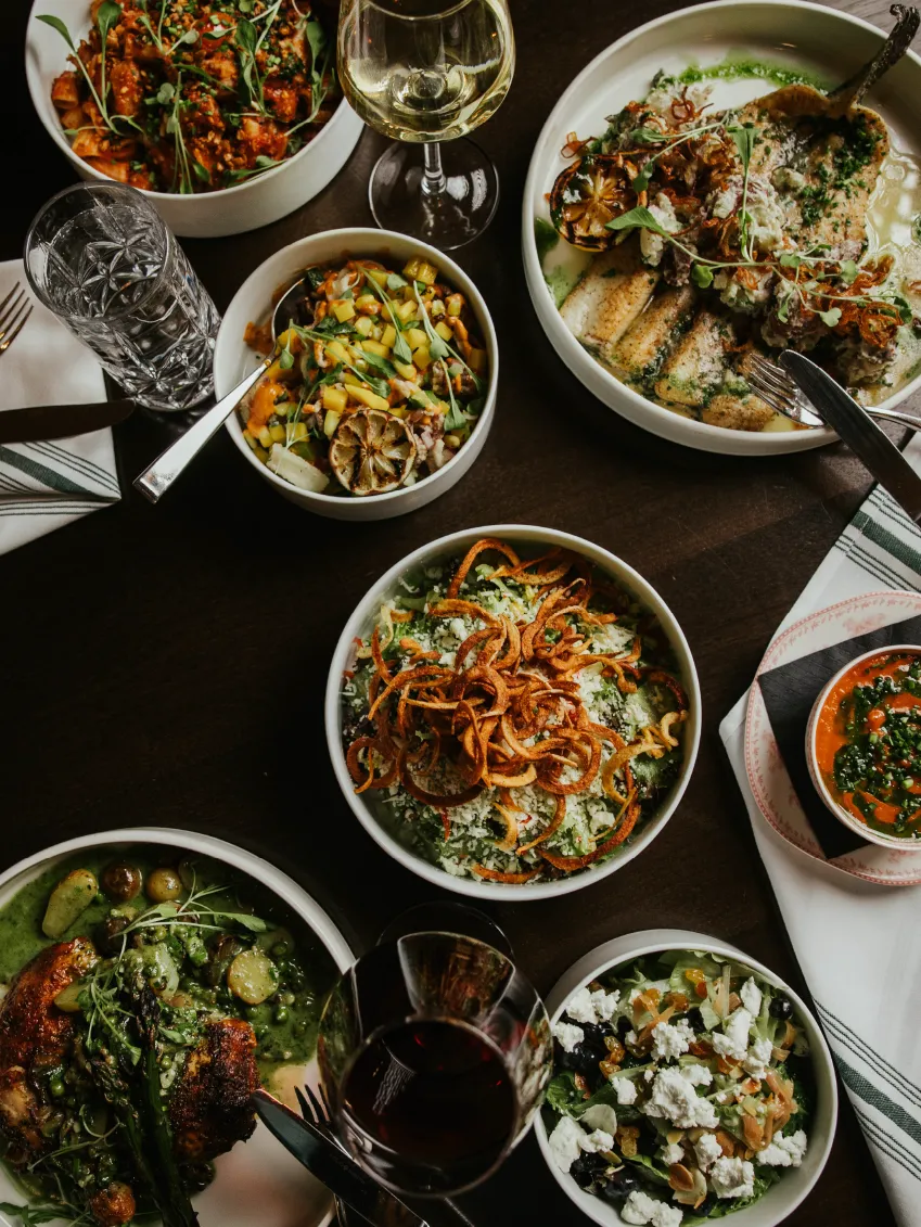 Table with assorted dishes including grilled fish, roasted chicken with green sauce, salads with greens and cheese, and two glasses of wine.