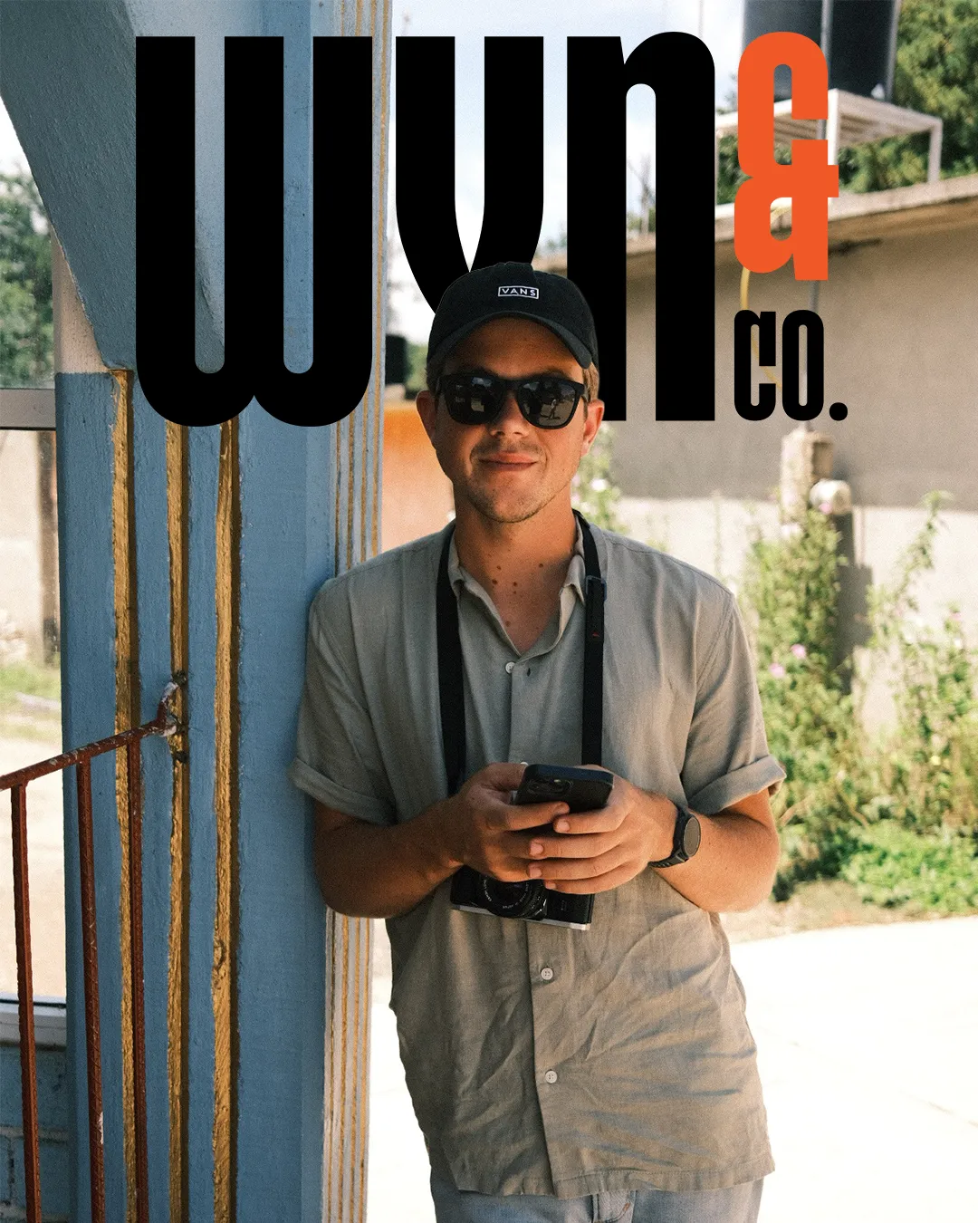 Man wearing sunglasses and a black Vans cap, holding a smartphone with a camera hanging from his neck, leaning against a painted blue wall outdoors.