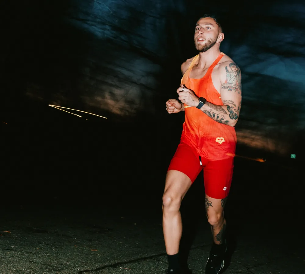Man with tattoos running outdoors at dusk wearing an orange tank top and red shorts.