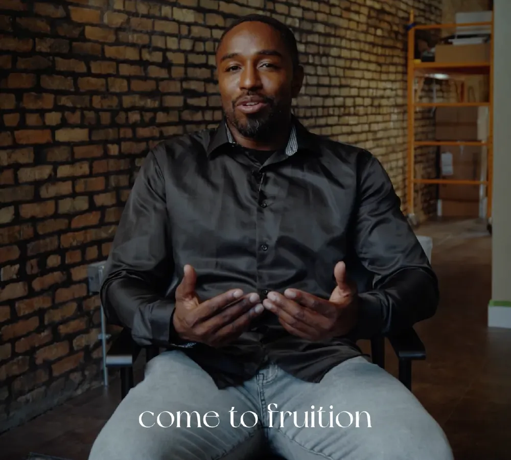 Man in black shirt sitting and speaking with a brick wall background and text 'come to fruition' at the bottom.