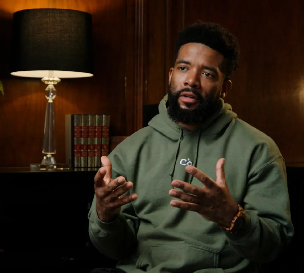 Bearded man in a green hoodie speaking with expressive hand gestures in a warmly lit room with a lamp and books behind him.
