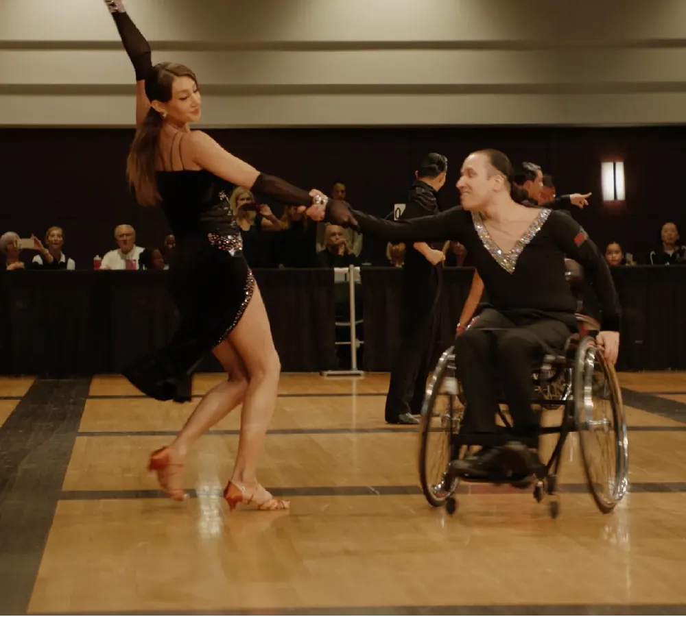 Man in wheelchair and woman in black dress dancing together on a wooden dance floor with an audience in the background.