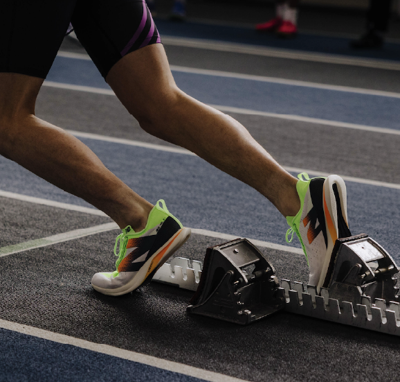 Athlete's legs and neon running shoes positioned on starting blocks on an indoor track.