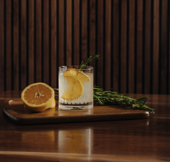 A glass of lemon drink garnished with lemon slices and a sprig of rosemary on a wooden board beside a halved lemon and rosemary sprigs.