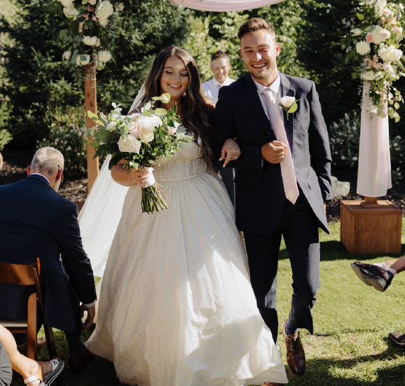 Bride in white gown holding bouquet and groom in dark suit walking arm-in-arm outdoors at a wedding ceremony.
