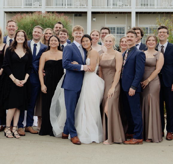 Bride and groom in wedding attire smiling and embracing, surrounded by bridesmaids and groomsmen dressed in formal dresses and suits outside a white building.